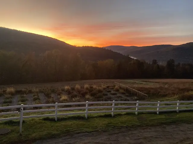 A vibrant orange and red sunset over rolling forested hills, viewed from the Hinata property with a white split-rail fence in the foreground