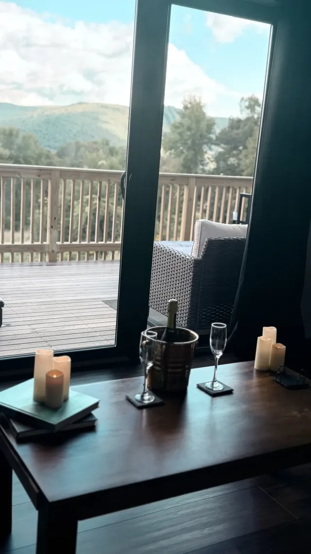 A candlelit table with wine glasses overlooks a wooden deck and mountain view.