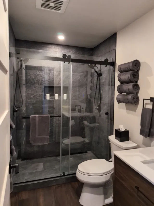 A modern bathroom with a glass-enclosed dark tile shower, white toilet, and gray towels.